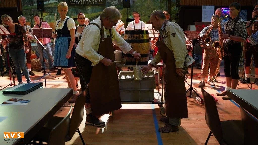 Zwei Männer in Schürzen zapfen ein Bierfass an, dahinter das Orchester in Tracht